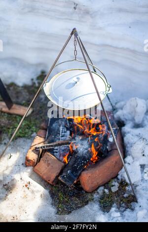 Over the fire hangs a pot in which to cook food. On a hook on a tripod, steam comes out of the pan. Winter Camping outdoor cooking Stock Photo