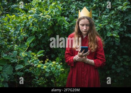 Preteen girl wearing red dress and paper crown standing in wet garden using phone Stock Photo