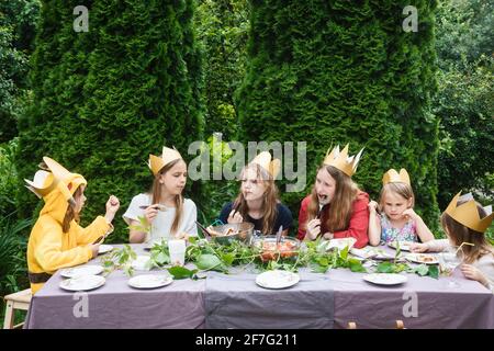 Children wearng paper crowns sitting by  decorated table eating grilled sausages celebrating birthday party in a green garden Stock Photo