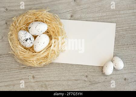 Easter eggs in a straw nest and greeting card over white wood. Template Easter greeting card. Top view with copy space. Stock Photo