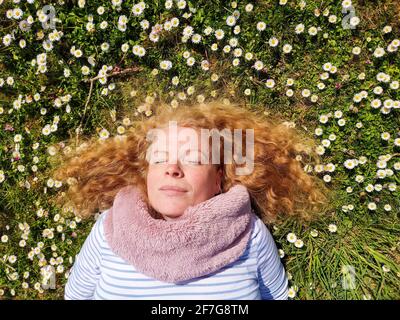 Beautiful redhead mature woman in her fifties lying in the grass in a field of daisies. Stock Photo