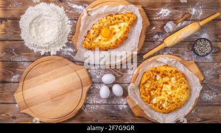 Khachapuri lies on wooden round pizza boards, chicken eggs and a rolling pin on a brown board background. Flat layout. Stock Photo