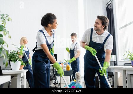 Interracial cleaners smiling at each other while holding mops in office Stock Photo