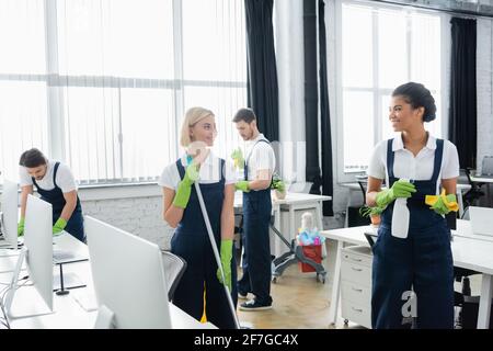 Interracial colleagues smiling at each other while holding cleaning supplies in office Stock Photo