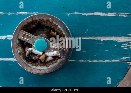 A retro, antique cigarette ashtray filled with stubs on a turquoise sanded wooden table top. Stock Photo