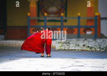 Kingdom of Bhutan, Asia, Himalaya. Two young novices in the courtyard of a monastery run hugging to the temple. Their red robes shine in the sun Stock Photo