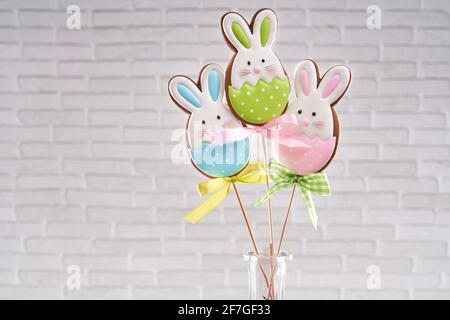 Hand painted gingerbread bunnies on wooden stick in glass vase. Tasty bright biscuits. Treats for children. Background of white brick wall Stock Photo