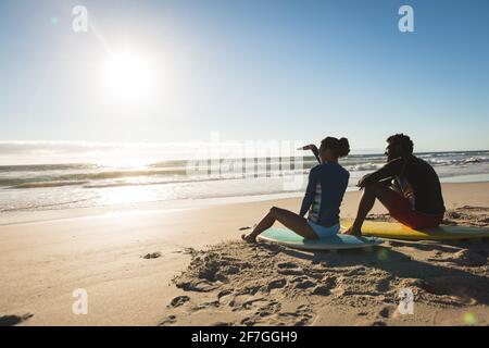 Happy african american couple on the beach sitting on surfboards looking toward sea Stock Photo