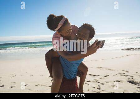 Happy african american couple on beach by the sea piggybacking and taking selfie Stock Photo