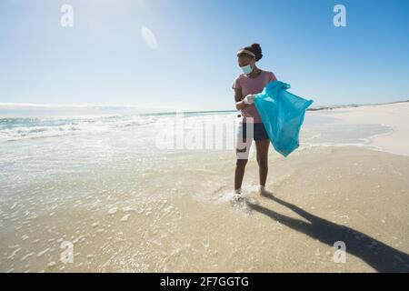 African american race woman wearing face mask collecting rubbish from the beach
