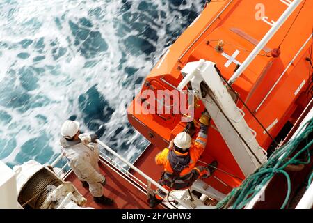 Crew member with orange jacket and helmet checking aft hook on life boat fixed to container vessel, second seaman in white overall observing. Stock Photo