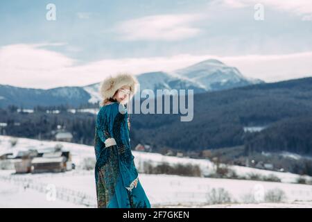 Young woman walks dressed in fur hat and dress against beautiful landscape of snowbound Carpathians mountains, sky and village.