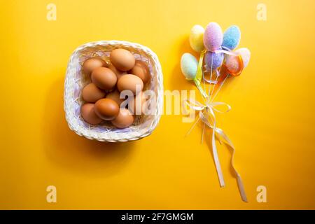 Basket with fresh eggs and fluffy colorful decorative plastic eggs isolated on yellow. Easter concept. Stock Photo