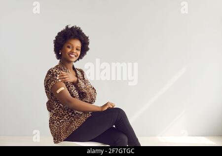 Portrait of happy woman sitting on chair and showing her arm after getting vaccinated Stock Photo