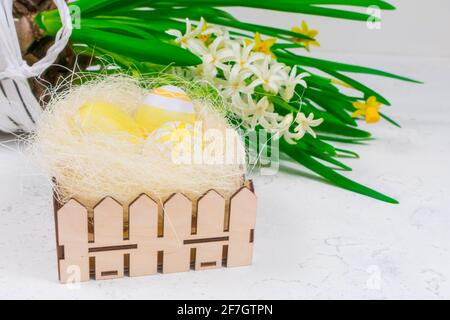 Easter concept. Yellow Easter eggs in a nest next to a bouquet of daffodils on a white concrete table. Place for an inscription. Base for coating. Stock Photo