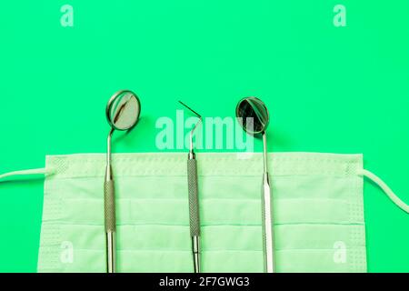 Dentist tools: mirror, dental explorer and tweezers lying on medical mask on neon green background. Stock Photo