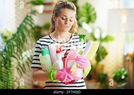 sad young female in striped shirt with cleaning supplies in the modern living room in sunny day. Stock Photo