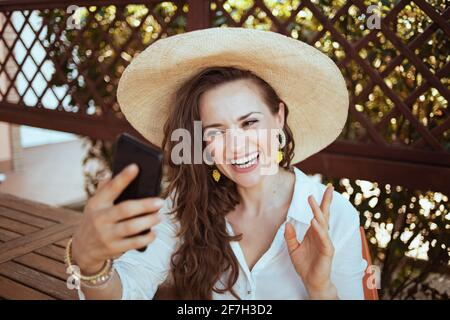 happy modern 40 years old woman in white shirt with hat having webcam meeting on a smartphone in the terrace of guest house hotel. Stock Photo