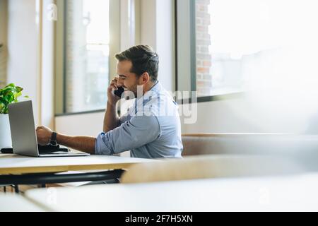 Young smiling man in office speaking on mobile phone. Male sitting in office with laptop using on smart phone. Stock Photo