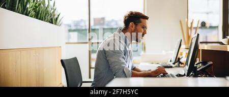 Freelancer looking at computer screen while typing. Young businessman sitting at desk looking at desktop monitor while typing on computer. Stock Photo