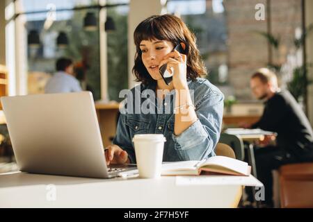 Young freelancer sitting in coworking space talking on cell phone and using laptop. Woman using laptop and speaking with client on mobile phone. Stock Photo