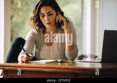 businesswoman making notes while talking on cell phone. Woman working from home. Stock Photo