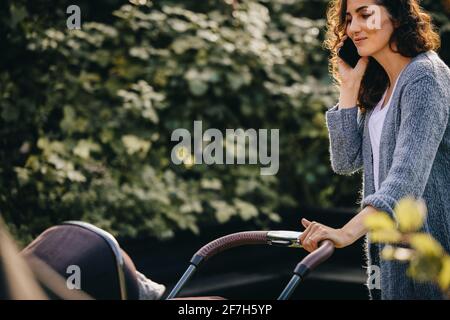 Mom walking around the neighbourhood pushing her baby in the stroller. Woman talking on mobile phone while pushing a pram. Stock Photo