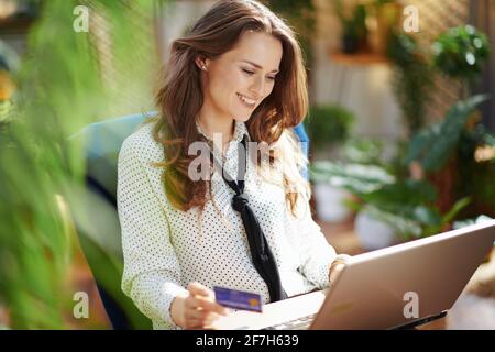 Green Home. smiling modern 40 years old housewife with long wavy hair with credit card using laptop in the modern living room in sunny day. Stock Photo
