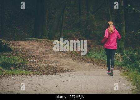 A female runner in black tights and pink jacket is running away from the camera on a gravel path towards the small bridge covered with fallen leaves. Stock Photo