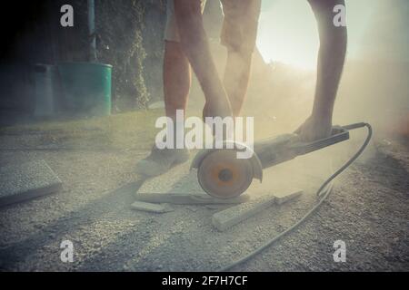 Man is cuttng a stone concrete tile on the floor using an electric angle grinder with dust and residue flying around on a sunny day outside. Stock Photo