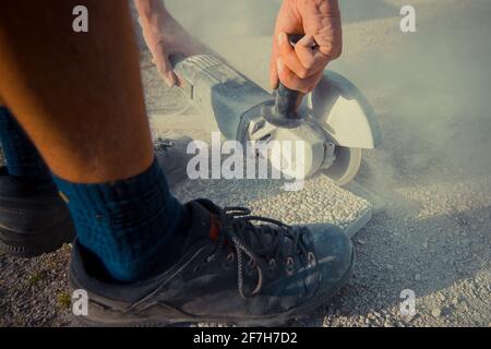 Man is cuttng a stone concrete tile on the floor using an electric angle grinder with dust and residue flying around on a sunny day outside. Stock Photo