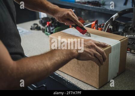 Hands of a man seen opening a brown cardboard box using a red utility knife. Service bench in the background. Concept of new parts in service. Stock Photo