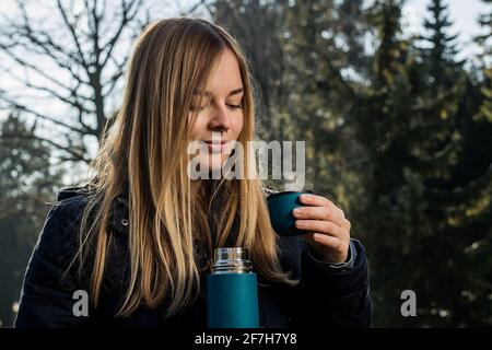 Cute blonde girl with long hair and septum piercing is drinking tea from a thermos mug in a park with some snow and strong sunny backlight. Looking to Stock Photo