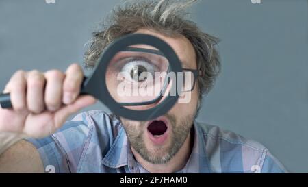 a curious funny surprised Middle-aged  man who is looking at the camera through a magnifying glass. Humorous portrait of a crazy man with no cut hair. Stock Photo