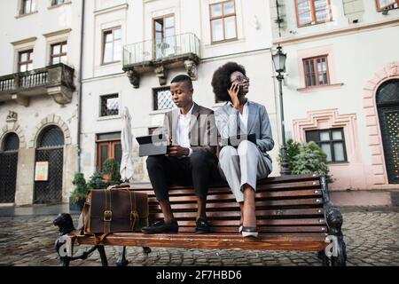 Handsome man in suit working on digital tablet while attractive woman talking on mobile and writing on clipboard. Two african colleagues solving working issues outside office. Stock Photo