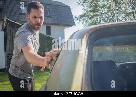 Young man with a hip beard cleaning a window of an old green vintage car on a home lawn. Cleaning muscle car windows at home with a cloth. Stock Photo