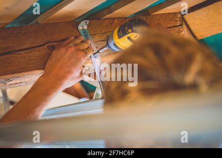 Screwing tin holders for a drywall into a wood portion of a roof using a battery powered cordless drill Stock Photo