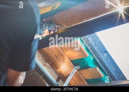 A person is cutting a board of wood next to a window with a reciprocating saw. Sawdust is flying around. Stock Photo