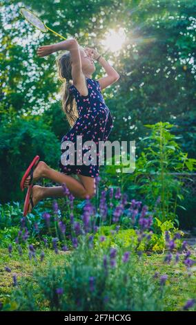 Young caucasian girl or kid is playing badminton game in a lush green garden. Kid jumping high up into sun while playing badminton Stock Photo