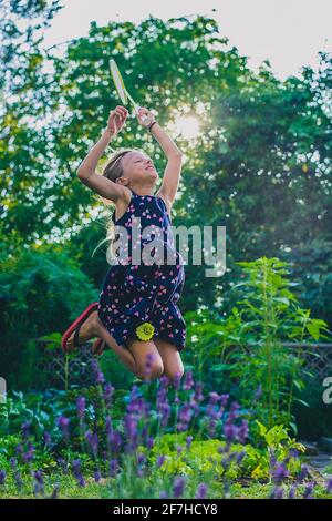 Young caucasian girl or kid is playing badminton game in a lush green garden. Kid jumping high up into sun while playing badminton Stock Photo