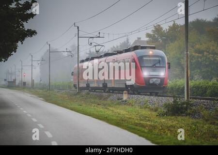 Early commuter train in slovenia in a foggy misty morning next to an adjacent road. Concept of faster and safe trains in regard to cars on roads. View Stock Photo