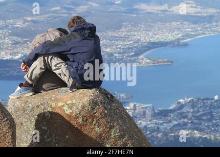 A couple sitting on a rock formation and enjoying the view over Hobart from the top of Mount Wellington, Hobart, Australia. Stock Photo