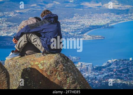 A couple sitting on a rock formation and enjoying the view over Hobart from the top of Mount Wellington, Hobart, Australia. Stock Photo