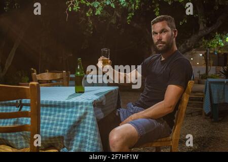 Young man is holding glass of beer in ambient of traditional greek tavern or restaurant at night while looking at camera. Table with checkered table c Stock Photo