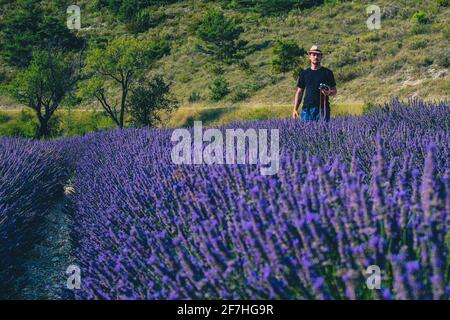 A young caucasian male with hipster image and thatched hat is stnading in a blooming lavender field with a vintage photo camera. Person looking away f Stock Photo