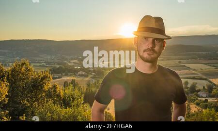 A young casual caucasian male with a thatched hat and black shirt is posing and looking into the camera in the evening hours with the sun just about t Stock Photo