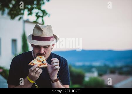 Hipster looking man with thatched hat  holding and eating a slice of pizza in evening with a french countryside in the background. Stock Photo