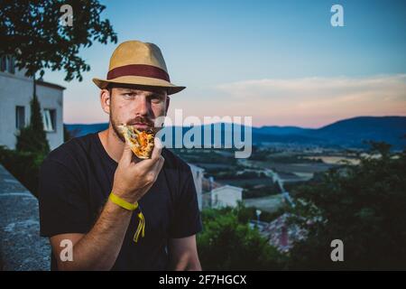 Hipster looking man with thatched hat  holding and eating a slice of pizza in evening with a french countryside in the background. Stock Photo