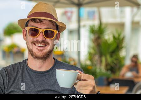 Hipster looking man with thatched hat and wooden sunglasses holding a white cup of coffee while drinking in smiling and sitting in a cafe environment. Stock Photo