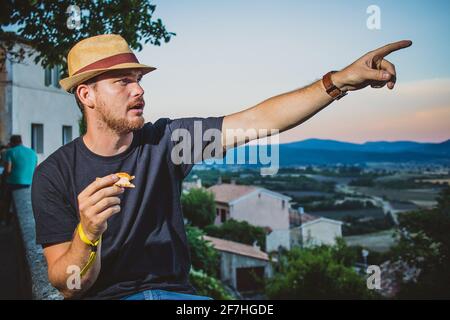 Hipster looking man with thatched hat  holding and eating a slice of pizza in evening with a french countryside in the background pointing with his ha Stock Photo